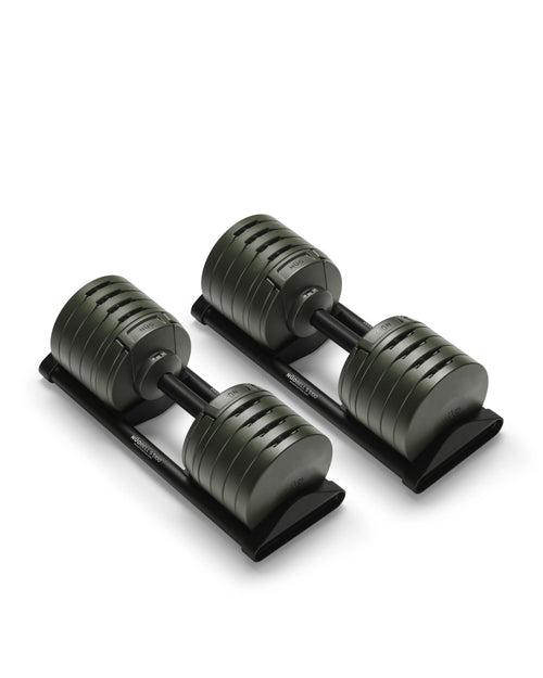 NÜOBELL S 5100 adjustable dumbbell pair in dark olive green finish positioned diagonally on black storage trays against white background, highlighting the thicker plate design with visible weight increment markings, the textured aluminum handle grip surface, and the precision-engineered stacking system that delivers 100 lb maximum capacity through the patented twist-handle selector.