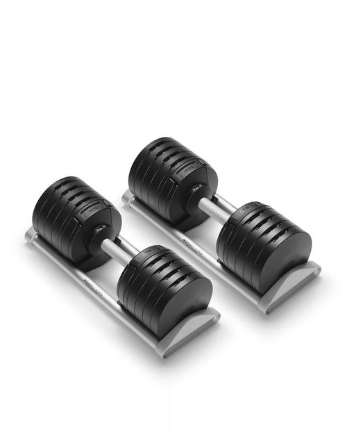 NÜOBELL S 5100 adjustable dumbbell pair in black finish positioned on brushed aluminum cradles, photographed at three-quarter angle showing the stacked weight plate architecture with visible increment markings, stainless steel V-connectors between plates, and the precision-machined twist selector mechanism that enables 5 lb adjustments across the full 5 to 100 lb range.