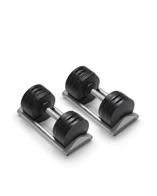 NÜOBELL S 560 adjustable dumbbell pair in matte black positioned at an angle on gray storage cradles, displaying the ergonomic 32mm aluminum handles with NÜOBELL S 560 branding, the layered four-plate construction with visible grooves between weight segments, and the flat-head end caps that create a stable base for resting between sets while maintaining instant access to 12 weight increments.