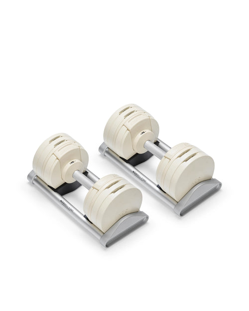 NÜOBELL S 560 adjustable dumbbell pair in cream finish resting on gray storage cradles with aluminum handles visible, showing the four-plate shifting system with stainless steel V-connectors and flat-head construction designed for stable racking and precise weight adjustments.